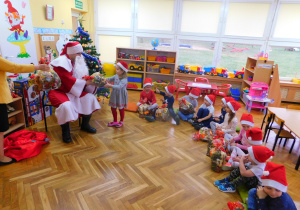 Dzieci siedzą z otrzymanymi paczkami, Mikołaj wręcza paczkę Zosi.