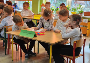 Szymon, Oliwia, Wiktoria i Zuzia siedzą przy stoliku i rozwiązują zadanie z treścią.