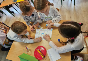 Szymon, Oliwia, Wiktoria i Zuzia siedzą przy stoliku i układają okienkowe puzzle.