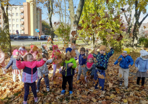 Dzieci na trawniku obrzucają się liśćmi.