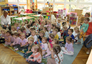 Dzieci siedzą na dywanie i podnoszą ręce do góry.