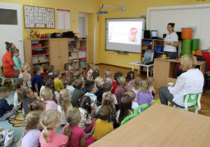 Dzieci z zaciekawieniem słuchają pani o oglądają obrazki na tablicy interaktywnej.
