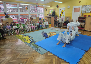 Dzieci siedzą i oglądają pokaz sztuki walki judo.