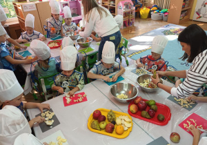 Pani Dyrektor i Pani Kamila obierają owoce a dzieci kroją je na cząstki.