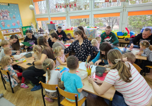 Rodzice wraz z dziećmi siedzą przy stolikach i wykonują plakaty