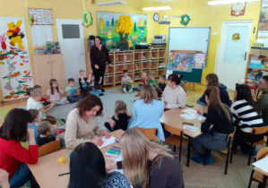 Rodzice pracują przy stolikach a dzieci siedzą na dywanie.