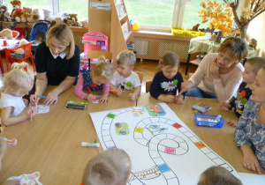 Dzieci przy stole kolorują obrazki i ozdabiają dużą grę planszową, pomaga mama Julki;