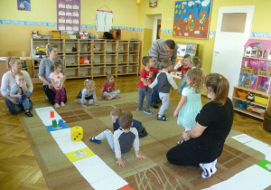 Grupa dzieci i rodziców gra w grę na dywanie, zespół z tatą Jagny wykonuje zadanie - taniec w kole;