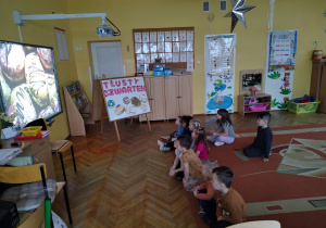 Dzieci siedzą na dywanie i oglądają film edukacyjny na temat tłustego czwartku.