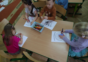 Dzieci siedzą przy stolikach i kolorują kolorowanki z pączkami.