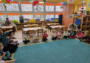 Dzieci naśladują jazdę na sankach w zabawie ruchowo-muzycznej, a przybyli goście obserwują