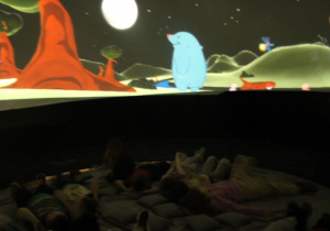 Dzieci wygodnie ułożone na podusiach oglądają film.