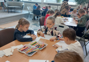 Przedszkolaki siedzą przy stolikach i kolorują ilustracje z kotami.