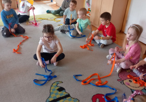 Dzieci siedzą na dywanie i trzymają patyczki ze wstążkami.