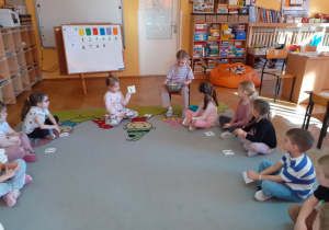 Dzieci siedzą w kole, Kaja wrzuca guziki do miski, Maja pokazuje kartonik z cyfrą.