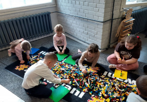 „Krasnoludki” siedzą po dwóch stronach dywanu z rozsypanymi klockami LEGO.