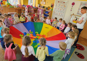 Dzieci trzymają chustę animacyjną a pan Damian wrzuca na nią krążki-owoce