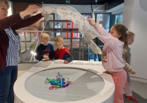 15.Lilia z panią Renatą sprawdzają działanie strumienia powietrza, unosząc lekkie elementy nad stanowiskiem eksperymentalnym, a Jakub i Leon regulują siłę nawiewu powietrza za pomocą pokręteł.