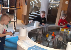 Antoś puszcza wodne obiekty i obserwuje, jak zmienia się ich tor w zależności od prądu, a Jakub P. i Jakub Cz. sterują przepływem wody, kierując strumień na kolorowe figurki w zbiornikach, aby sprawdzić siłę i zachowanie wody.