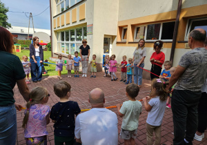 Panie wraz z dziećmi, rodzicami i dziadkami trzymają gumę sensoryczną i słuchają zasad zabawy, które objaśnia pani Kamilka