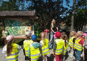Pani przewodnik stoi i opowiada zebranym dzieciom o powstaniu MiniZoo.
