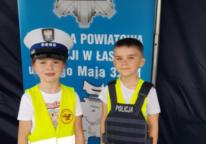 Michalina w policyjnej czapce i Kacper w kamizelce ochronnej pozują na tle policyjnej odznaki.