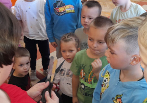 Pani Klaudia prezentuje dzieciom specjalistyczne narzędzia wykorzystywane do wyrównywania i dekorowania ciast, zachowując przy tym zasady bezpieczeństwa.