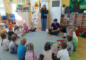 Dzieci siedzą na dywanie i słuchają objaśnień prowadzącej.