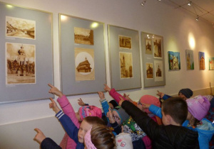  Korytarz biblioteki, na ścianie obrazy z Łaskiem, grupa dzieci ogląda i pokazuje obraz z kolegiatą;