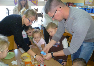  Mama Olka i tata Filipa pomagają dzieciom założyć balon na butelkę z roztworem z drożdży.
