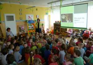 Dzieci siedzą przed tablicą interaktywną, prowadząca omawia autorów literatury dla dzieci.