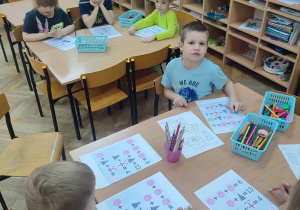 Dzieci i przy stolikach rozwiązują zagadki matematyczne w kartach pracy.