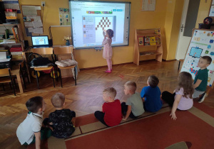 Dzieci siedzą na dywanie. Oliwka przy tablicy interaktywnej rozwiązuje zadania z kodowania.