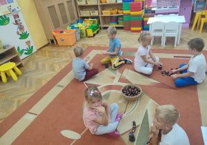 Dzieci na dywanie rozwiązują zagadki i zadania z kubeczkami i kasztanami.