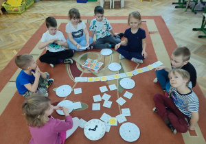 Dzieci na dywanie grają w grę. Szukają zegara z odpowiednią godziną.