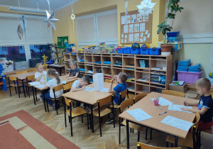 Dzieci siedzą przy stolikach i wykonują zadania w kartach pracy.