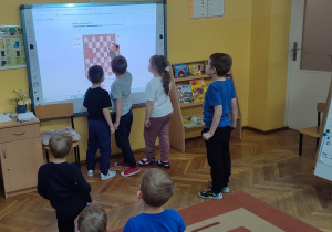 Dzieci stoją przy tablicy interaktywnej i rozwiązują zadania szachowe.