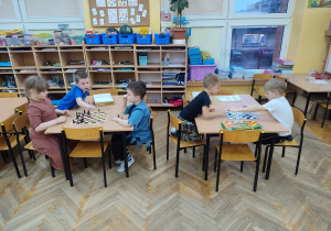 Dzieci siedzą przy stolikach i grają w szachy i w warcaby.