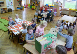 Dzieci podzielone na 3 zespoły: przy jednym stoliku malują farbami 2 domy z pudeł, przy drugim przyklejają „dachówki” na kartonie, przy trzecim – kolorują sylwety okien i drzwi;