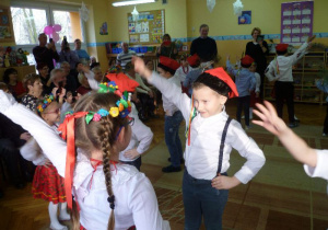 Dzieci parami w strojach krakowskich stoją w parach z ręką w górze - tańczą Krakowiaka, widoczna część widowni