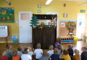 Dzieci siedzą przed teatrzykiem, oglądają przedstawienie „Pluszowy Miś odwiedza wujka Niedźwiedzia” (w wykonaniu nauczycielki).