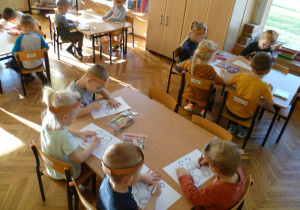 Dzieci przy stolikach kolorują kredkami obrazki przedstawiające różne misie.