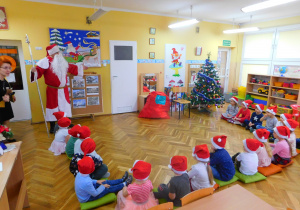 Dzieci siedzą w kręgu i witają wchodzącego do sali Mikołaja.