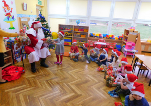 Dzieci siedzą z otrzymanymi paczkami, Mikołaj wręcza paczkę Zosi.