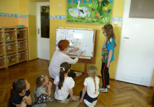  Dzieci siedzą pod tablicą, Ilonka z pomocą nauczycielki rozszyfrowuje hasła do puzzli z godłem;
