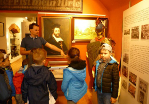  Dzieci stoją w sali muzealnej przed portretem Jana Łaskiego i słuchają przewodnika