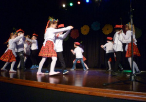 12 dzieci w strojach ludowych, ustawionych w parach na scenie tańczy krakowiaka