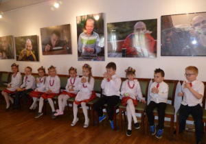 10 dzieci siedzi z poczęstunkiem pod ścianą z dużymi zdjęciami w "Sali z fortepianem";