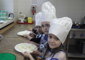 Weronika, Adaś, Sara przekazują syrnikowe placuszki do smażenia w przedszkolnej kuchni.