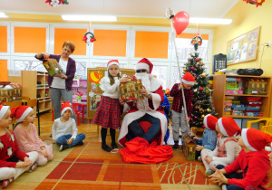 Mikołaj wręcza paczkę Sarze, a pani Renia pomaga odczytać imiona kolejnych dzieci.